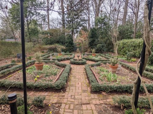 A serene garden with neatly arranged flower beds, stone pathways, and potted plants surrounded by trees.