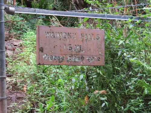 Sign for Waimoku Falls, indicating it's 1.5 miles away, with a note to please close the gate. Surrounded by greenery.