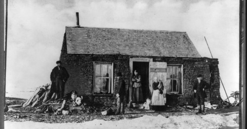 A historic black-and-white photo of a small stone house with five people standing outside in a snowy landscape.