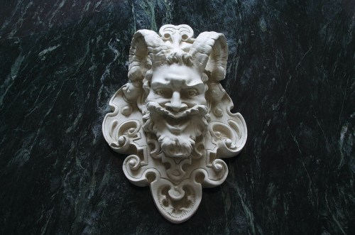 A white marble relief of a bearded face with horns, set against a dark green marble background.