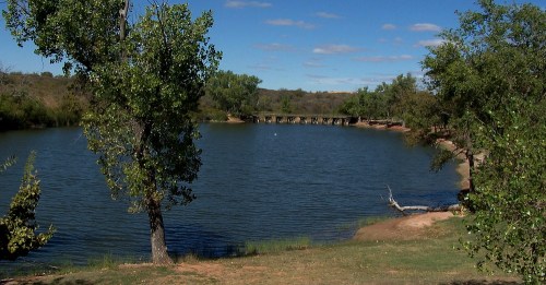 A serene lake surrounded by trees, with a clear blue sky and a distant bridge in the background.