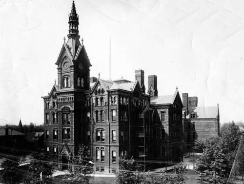 Historic brick building with a tall clock tower, surrounded by trees and other structures, in black and white.