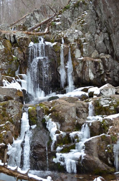 A cascading waterfall with icicles, surrounded by rocky terrain and patches of snow.