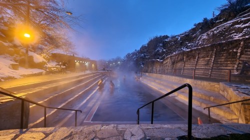 A serene hot spring surrounded by snow, with steam rising and people enjoying the warm water at dusk.