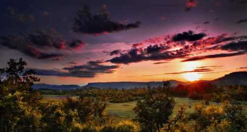 A vibrant sunset over a landscape with rolling hills, silhouetted trees, and colorful clouds in the sky.