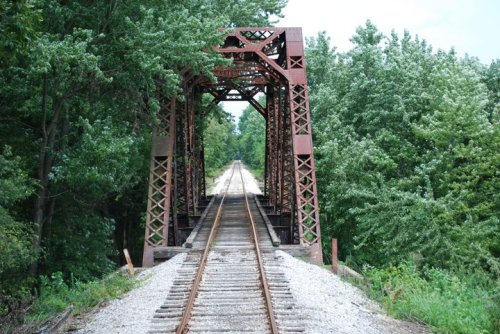 A rusty metal bridge spans a tree-lined path, with railway tracks running down the center.