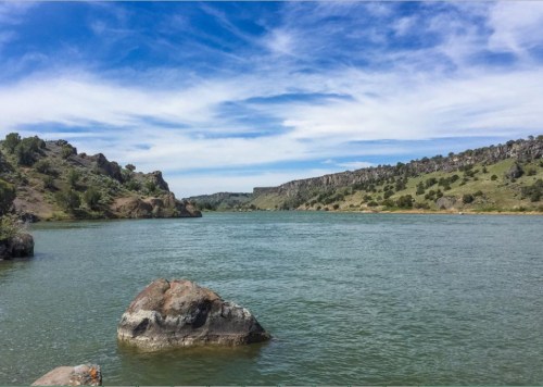 A serene river flows between rocky banks under a blue sky with scattered clouds. Lush greenery lines the shore.
