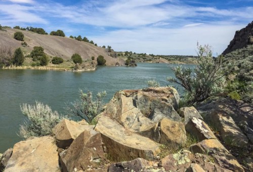 A serene landscape featuring a river surrounded by rocky shores and rolling hills under a blue sky with clouds.
