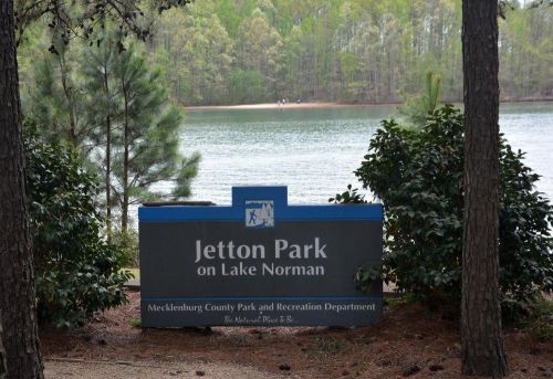 Sign for Jetton Park on Lake Norman, surrounded by trees and greenery, with a lake visible in the background.