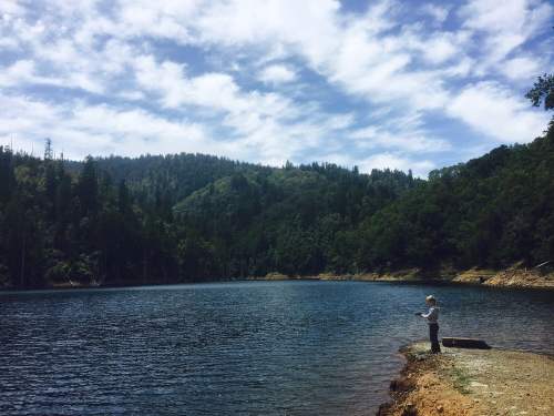 A person stands by a serene lake surrounded by lush green hills and a partly cloudy sky.