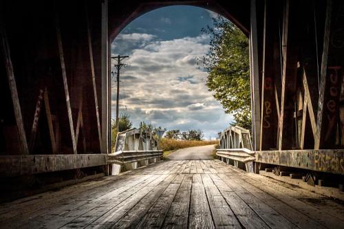 View from inside a covered bridge, leading to a winding road under a cloudy sky, surrounded by trees.