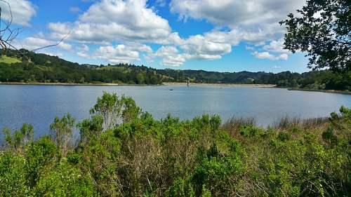 A serene lake surrounded by greenery and hills, under a partly cloudy sky.