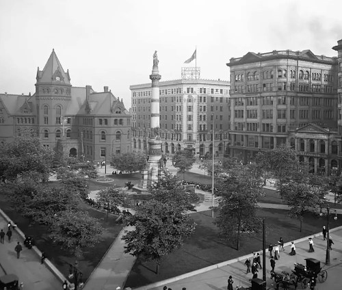 Historic black-and-white view of a city square with a monument, buildings, and trees, bustling with people and vintage cars.