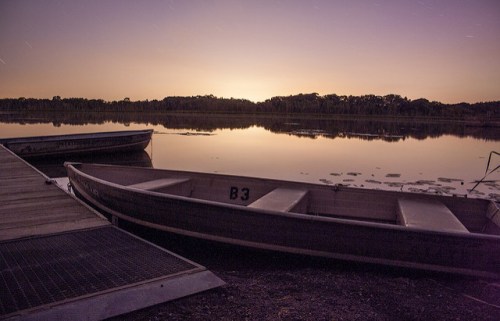 A serene lake at dusk with two boats docked and a colorful sunset reflecting on the water.