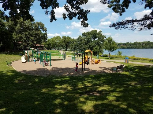 A playground with colorful equipment near a lake, surrounded by trees and grassy areas under a blue sky.