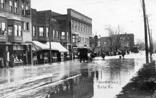 Historic black-and-white photo of a flooded street with buildings, people, and a horse-drawn carriage.