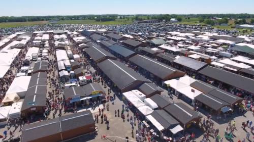 Aerial view of a bustling outdoor market with numerous stalls and a large crowd, surrounded by green fields and parking.