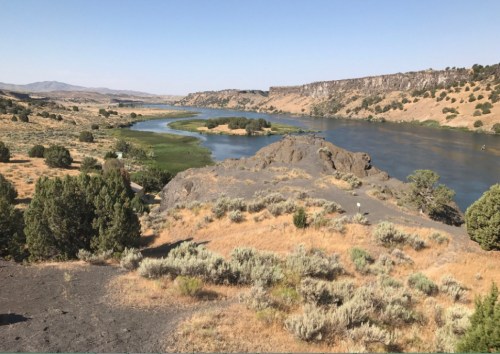 A scenic view of a river winding through a dry landscape with shrubs and rocky terrain under a clear blue sky.