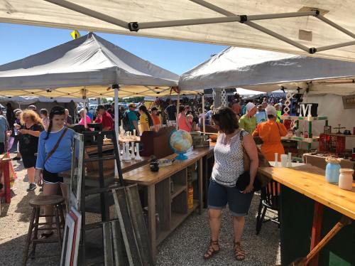 A bustling outdoor market with people browsing various stalls under white tents, showcasing antiques and crafts.
