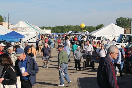 A busy outdoor market scene with people walking among tents and vendors on a sunny day.