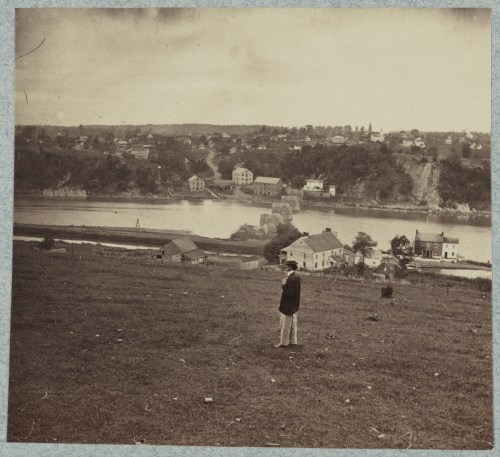 A sepia-toned photo of a man standing on a hill, overlooking a river and a small town in the distance.