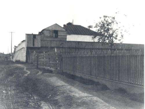 Black and white image of a wooden building with a fence and dirt path, surrounded by trees and power lines.