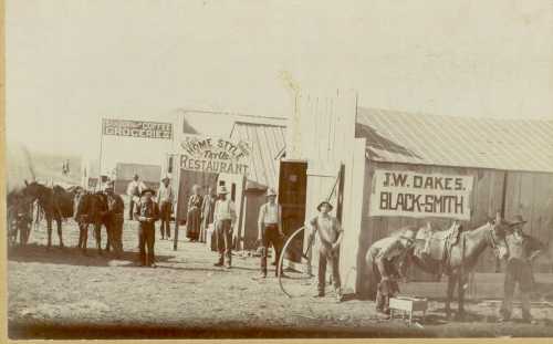 Historic photo of a western town with men, horses, and buildings for a grocery, restaurant, and blacksmith shop.