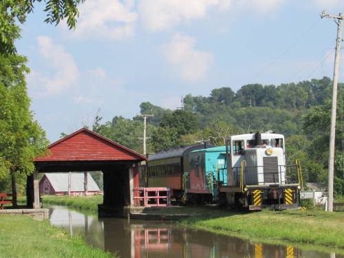 A colorful train parked beside a covered bridge, reflecting in a calm waterway, with green hills in the background.