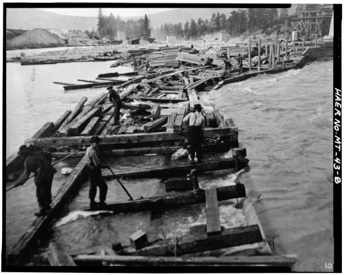 Workers on a wooden dock, surrounded by logs and water, with a forested area in the background. Black and white photo.