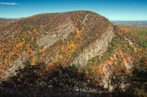 A scenic view of a mountain covered in autumn foliage, showcasing vibrant orange and green trees against a clear sky.