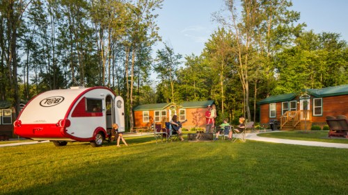 A cozy campsite with a retro camper, people gathered around a fire, and wooden cabins surrounded by trees.
