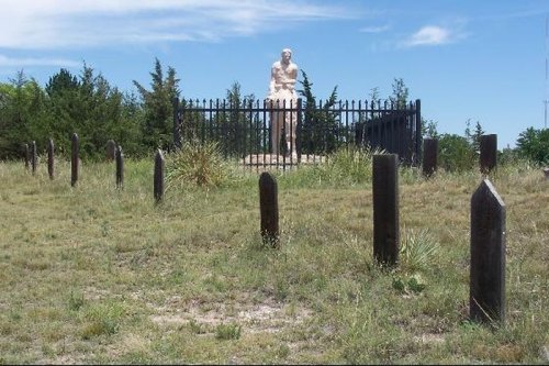 A statue surrounded by a black fence, with wooden markers in a grassy area under a blue sky.