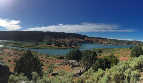 A panoramic view of a serene landscape featuring a river, green vegetation, and distant hills under a clear blue sky.