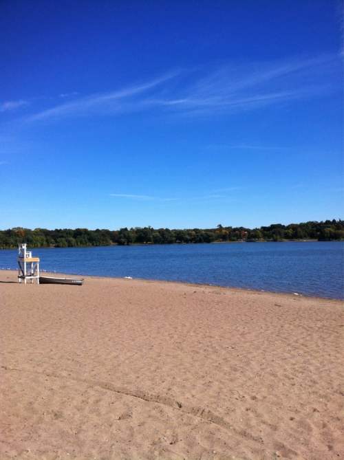 A sandy beach with a calm lake and clear blue sky in the background, surrounded by green trees.