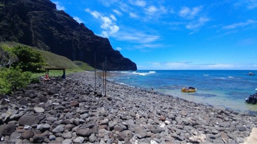 A rocky shoreline with clear blue water, cliffs in the background, and a small boat near the shore under a bright sky.