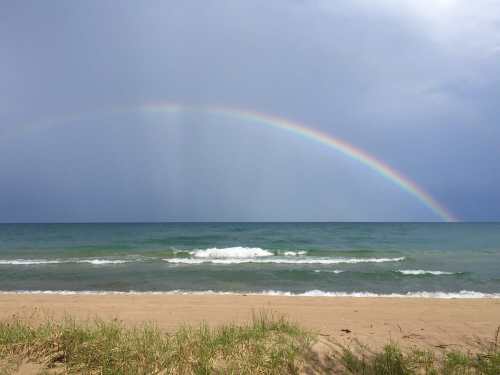 A vibrant rainbow arcs over a calm ocean, with gentle waves lapping at a sandy beach under a cloudy sky.