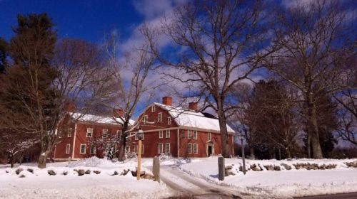 A red historic house surrounded by snow, with bare trees and a blue sky in the background.