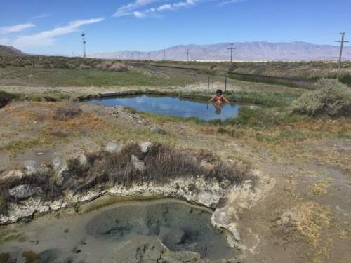 A person relaxes in a natural hot spring surrounded by grassy terrain and distant mountains under a clear blue sky.