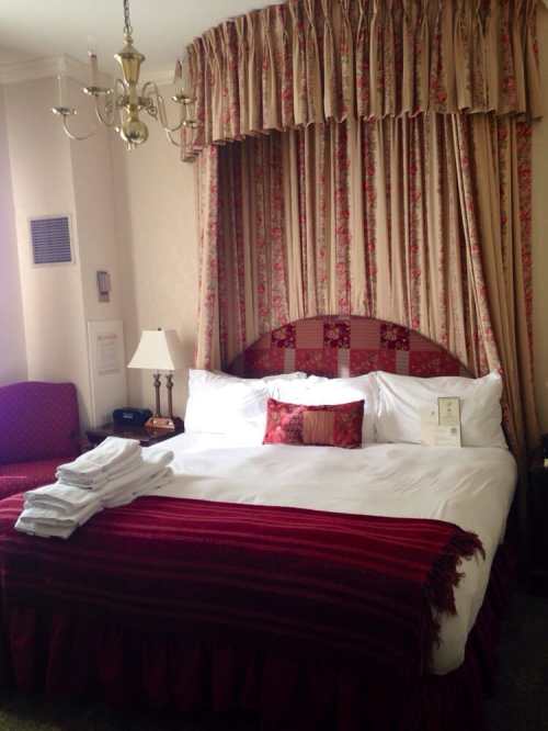 Cozy hotel room featuring a bed with red and white bedding, floral curtains, a lamp, and a plush chair.