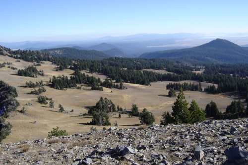 A panoramic view of a mountainous landscape with rolling hills, scattered trees, and a clear blue sky.