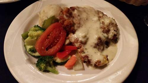 A plate of fried chicken topped with white gravy, served with mashed potatoes and a side of mixed vegetables and tomato.