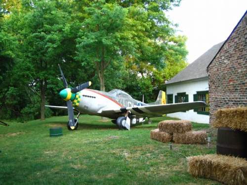 A vintage airplane parked on grass near a barn, surrounded by trees and hay bales.