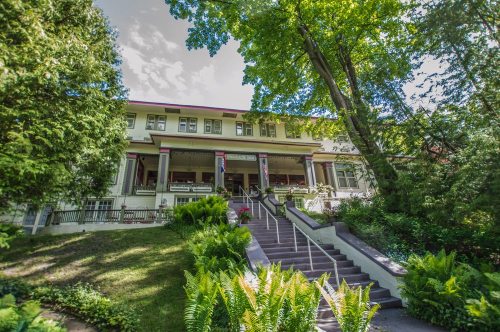 A large, historic house with a porch, surrounded by greenery and trees, featuring stairs leading up to the entrance.