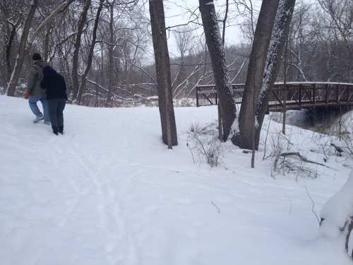 Two people walk on a snowy path near a river, with trees and a wooden bridge in the background.