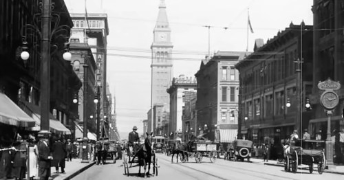 Historic street scene with horse-drawn carriages, buildings, and a clock tower in the background. Black and white photo.