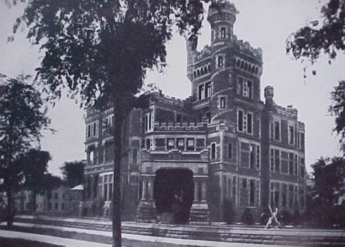 Historic black-and-white photo of a large, ornate castle-like building surrounded by trees.