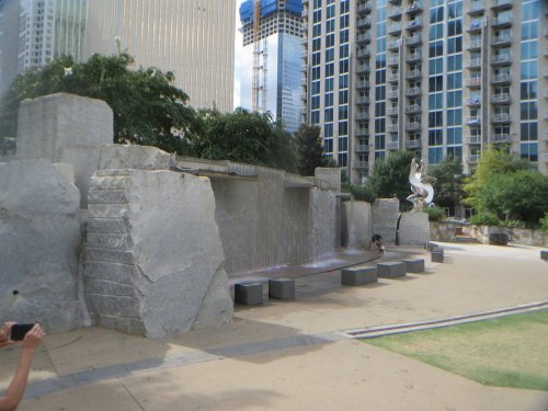 A modern water feature with a stone wall and cascading water, surrounded by greenery and tall buildings in the background.