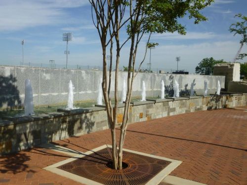 A tree stands near a stone fountain with multiple water jets, set against a blue sky and sports field in the background.