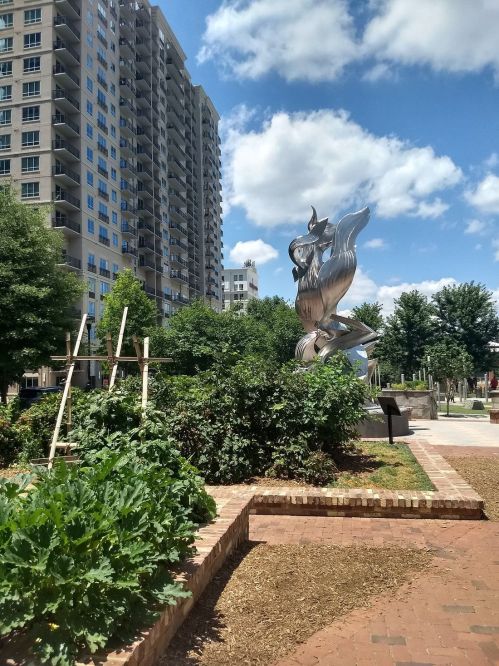 A shiny silver sculpture amidst greenery and urban buildings under a blue sky with fluffy clouds.