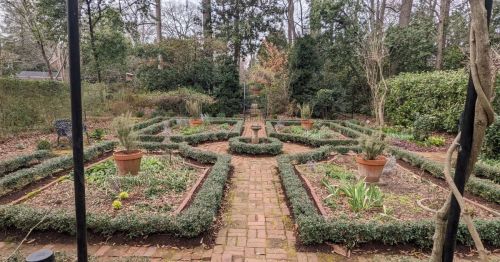 A serene garden with neatly arranged flower beds, brick pathways, and potted plants surrounded by lush greenery.